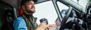 Young truck driver drinking from a coffee mug in the cab of his truck.